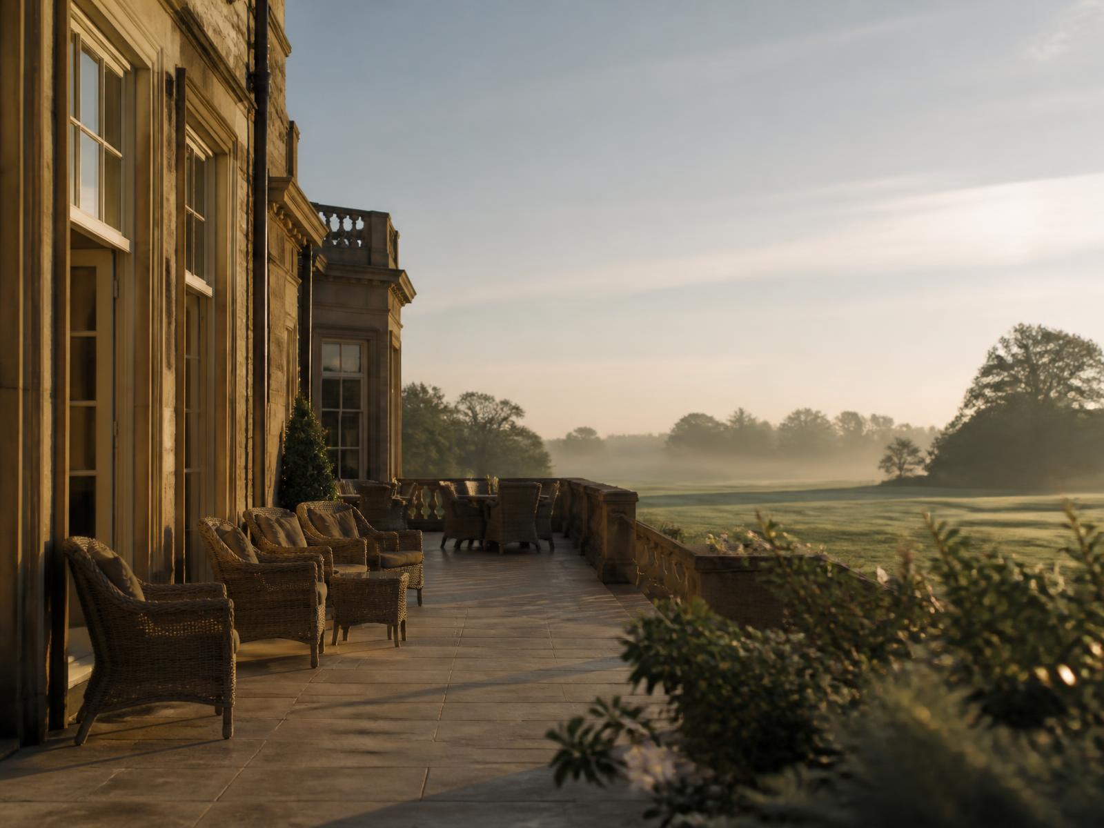 Empty terrace of a stone private golf clubhouse at sunrise, wicker chairs facing a misty fairway